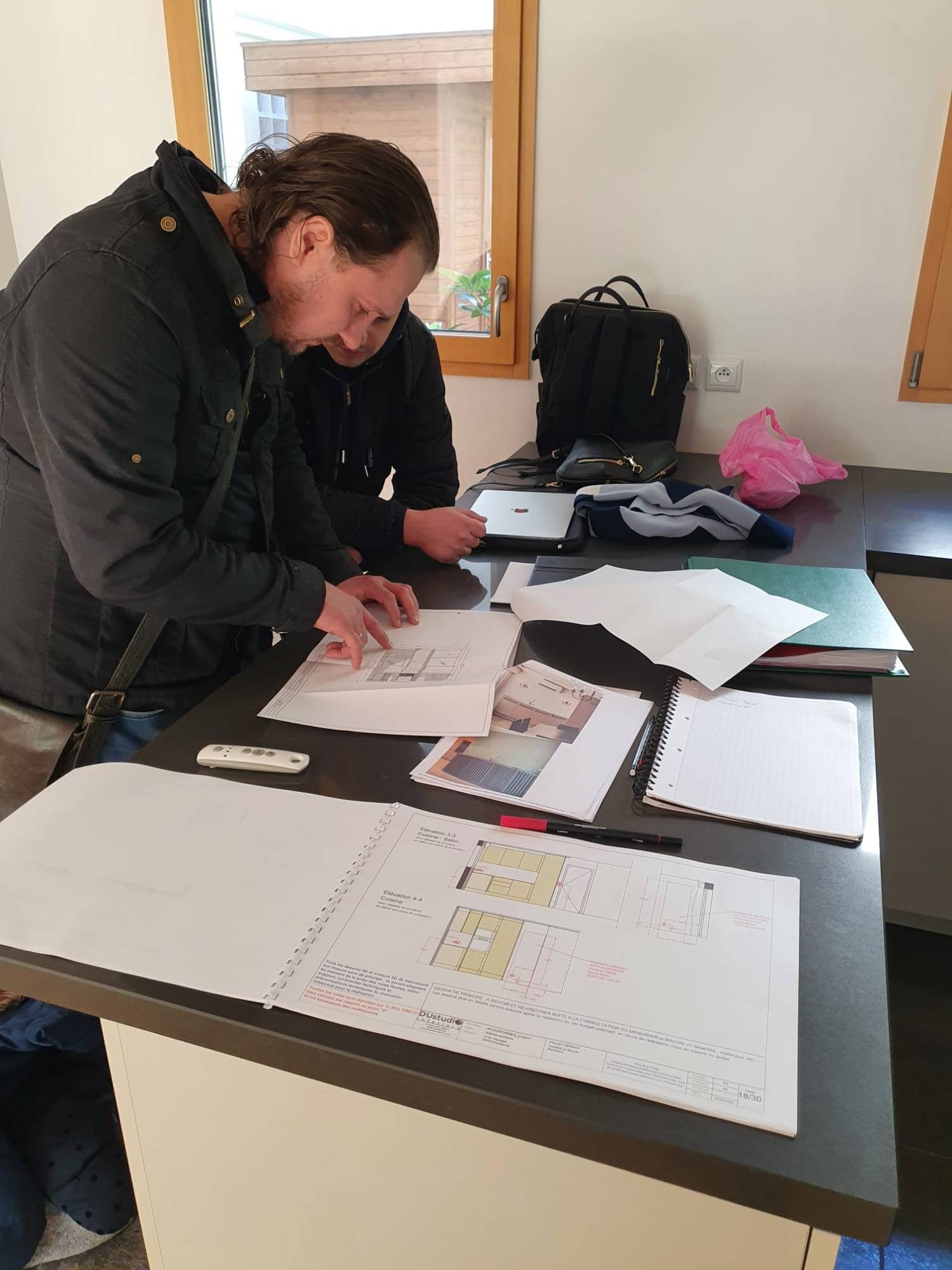 Deux personnes penchées sur des plans d'élévation de cuisine étalés sur un plan de travail, avec carnet de notes et dossiers.