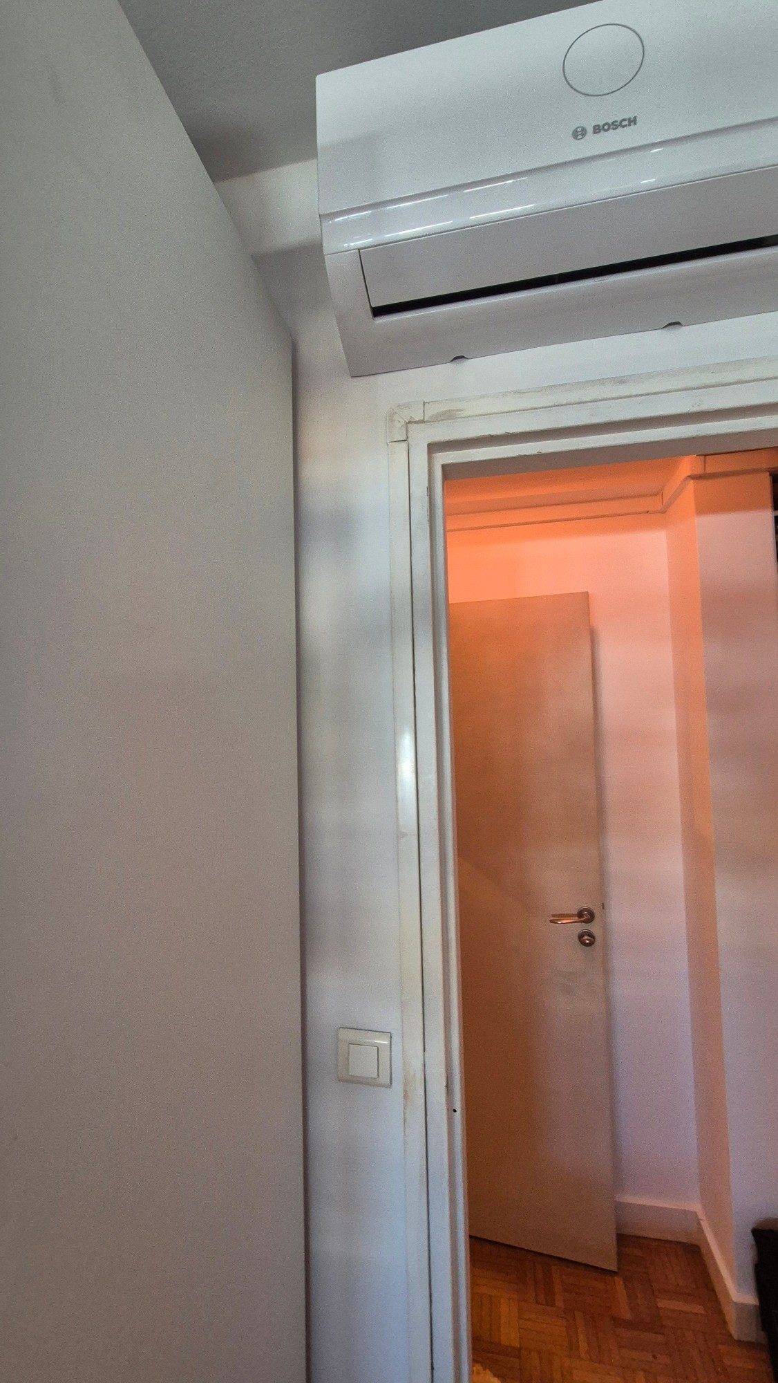Unité intérieure murale de climatisation réversible posée en applique au-dessus de l'encadrement d'une porte, dans une chambre aux murs clairs.