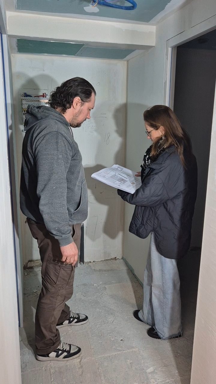 Deux personnes consultant un carnet de plans devant un tableau électrique en cours d'installation, dans un couloir en travaux.